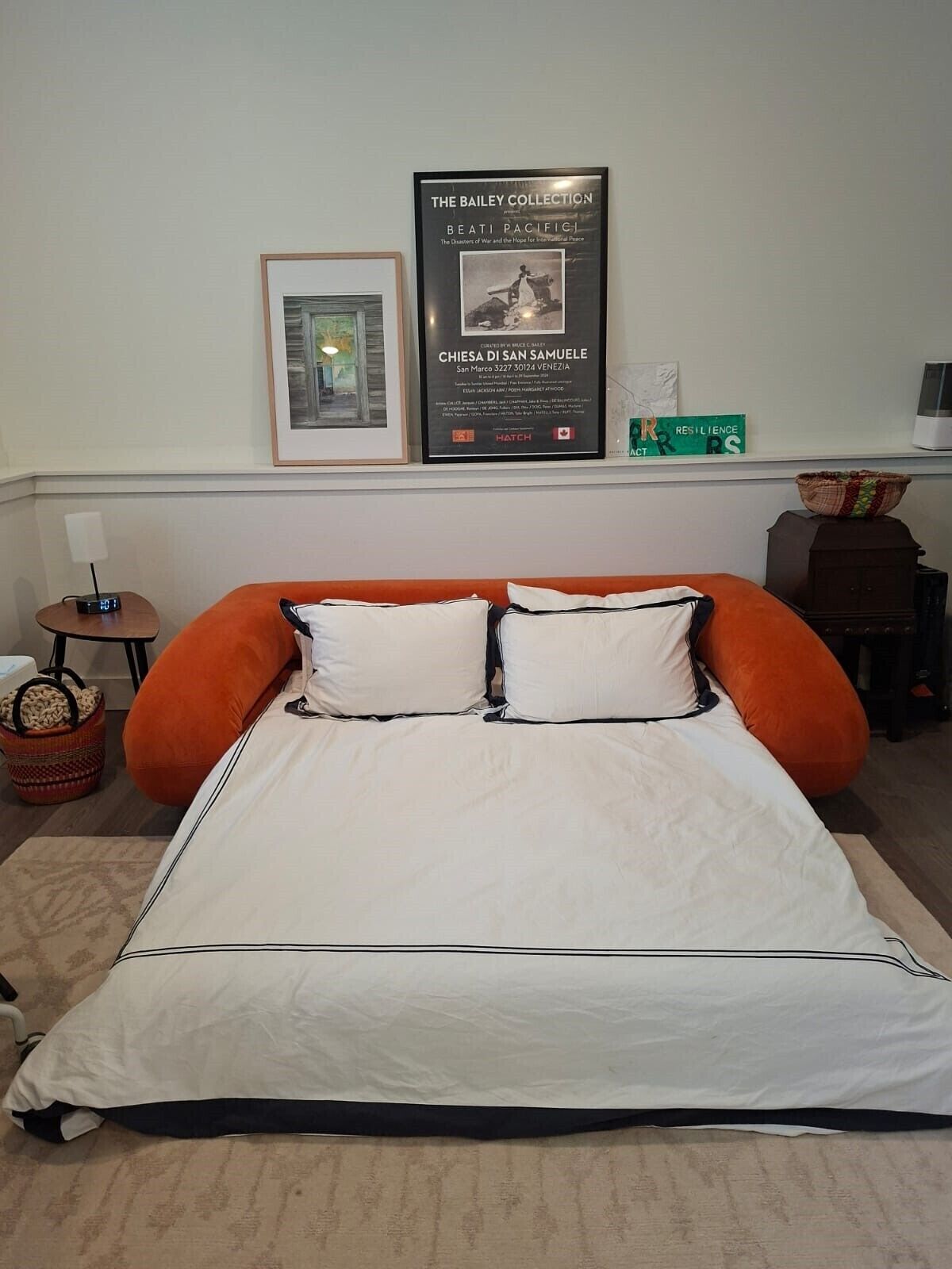 A hotel room with a large bed, a framed picture on the wall, a small table with a lamp, and a basket on the floor.