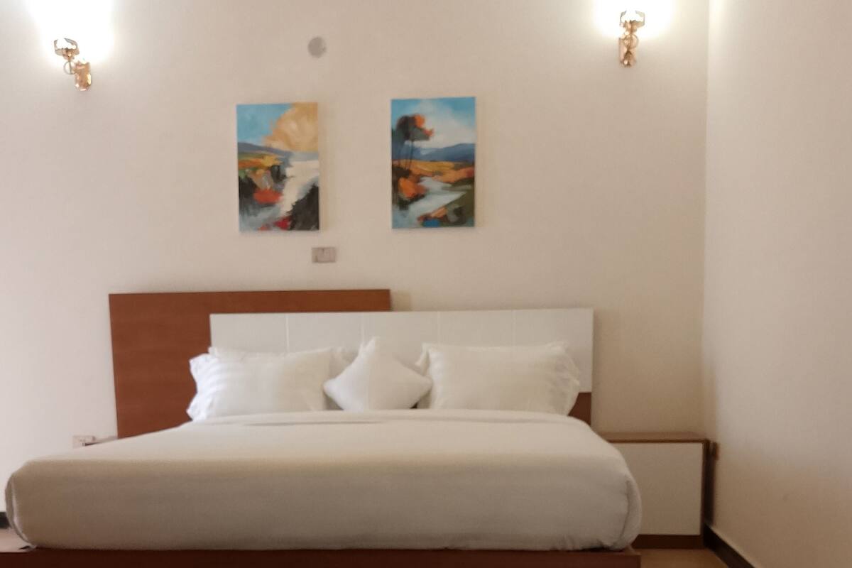 A bedroom with a wooden platform bed, white bedding, two wall-mounted paintings, and a black rug.