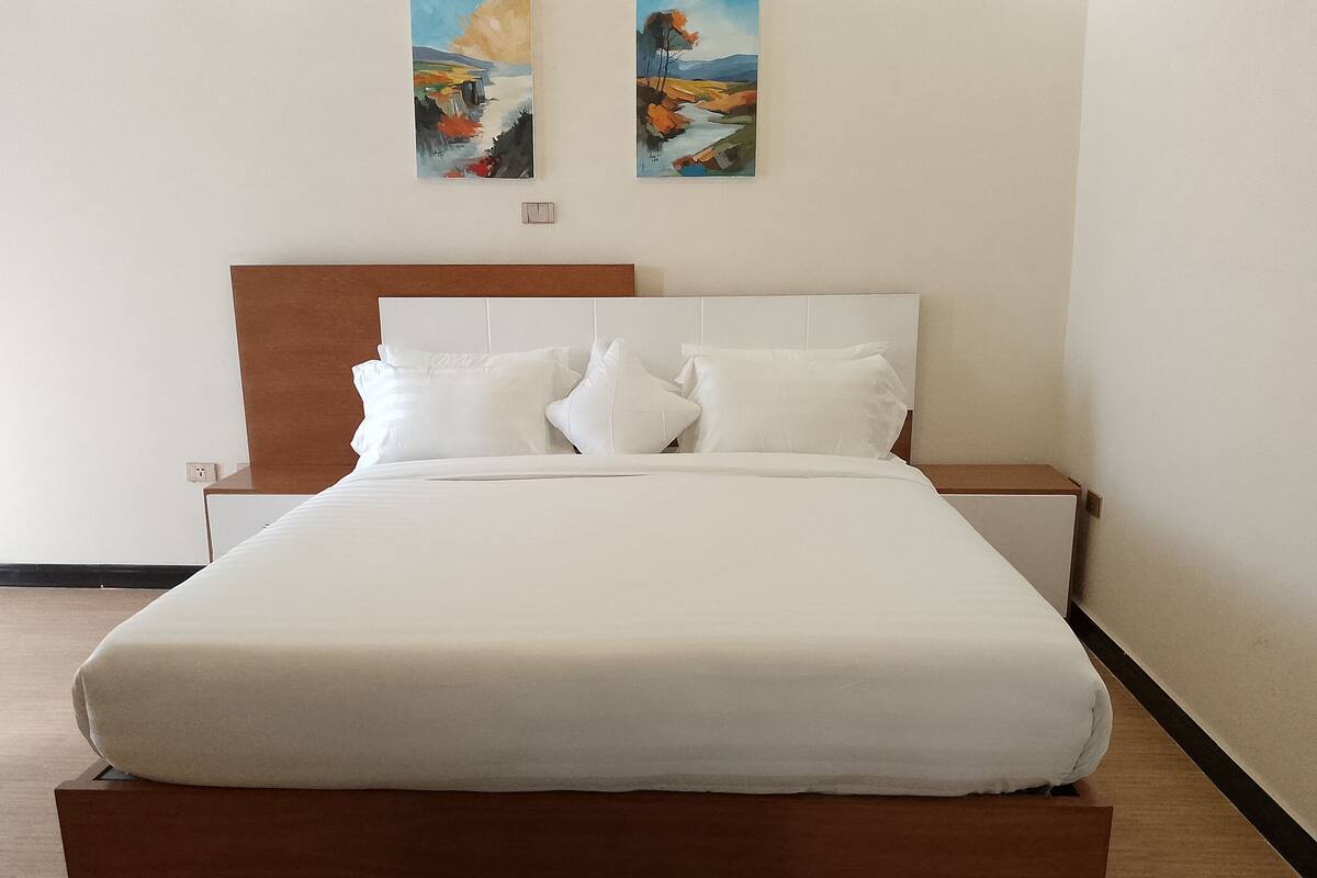 A neatly made bed with white linens and pillows, a wooden headboard, and two paintings on the wall.