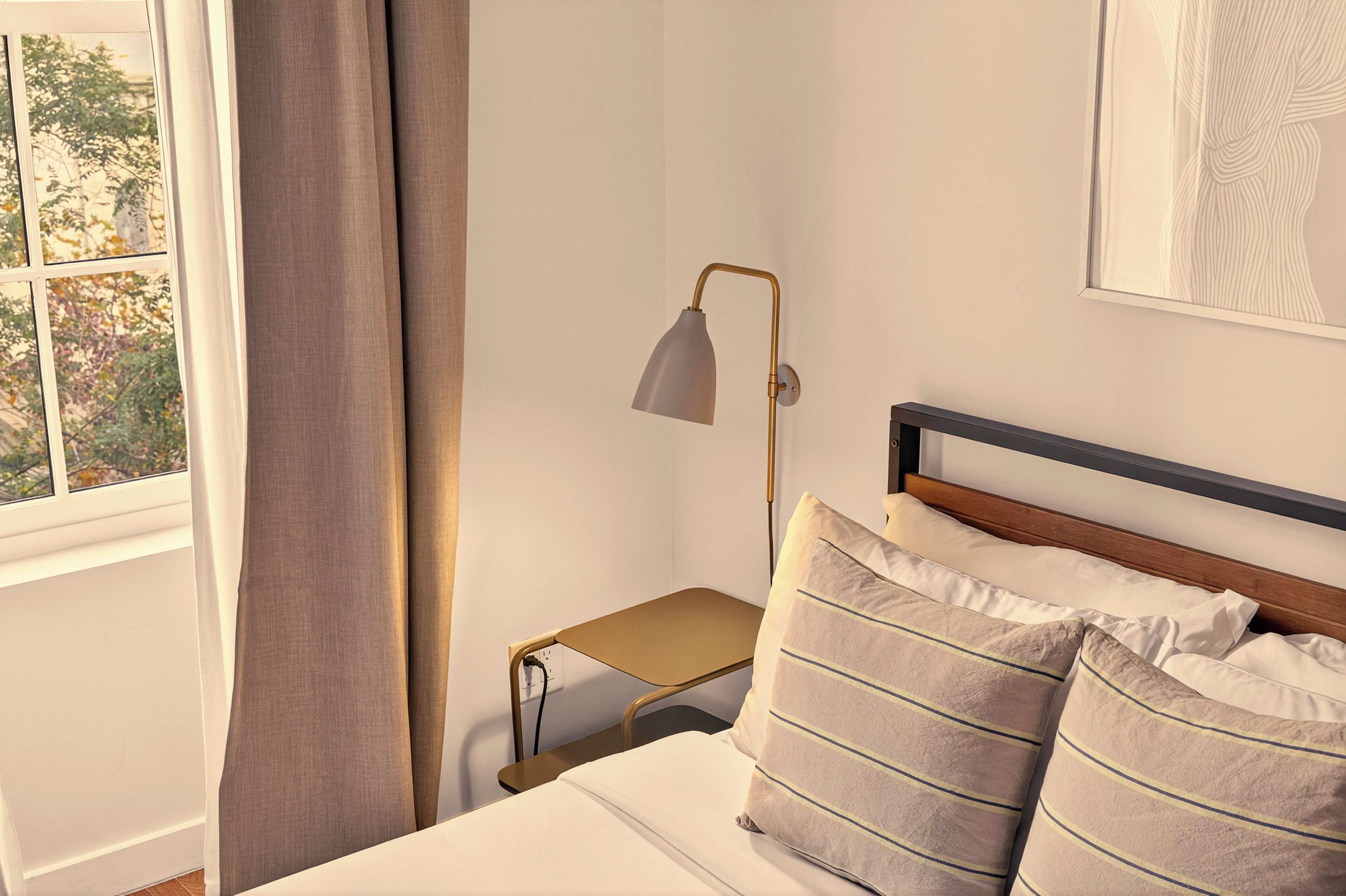 Una cama bien tendida con almohadas, una mesita de noche con lámpara y una ventana con cortinas.