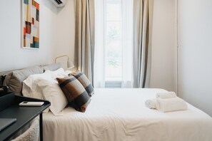 A neatly made bed with white linens, a laptop on a side table, and a wall-mounted air conditioner. - Outsite Chelsea - Work, Connect, Explore (New York)