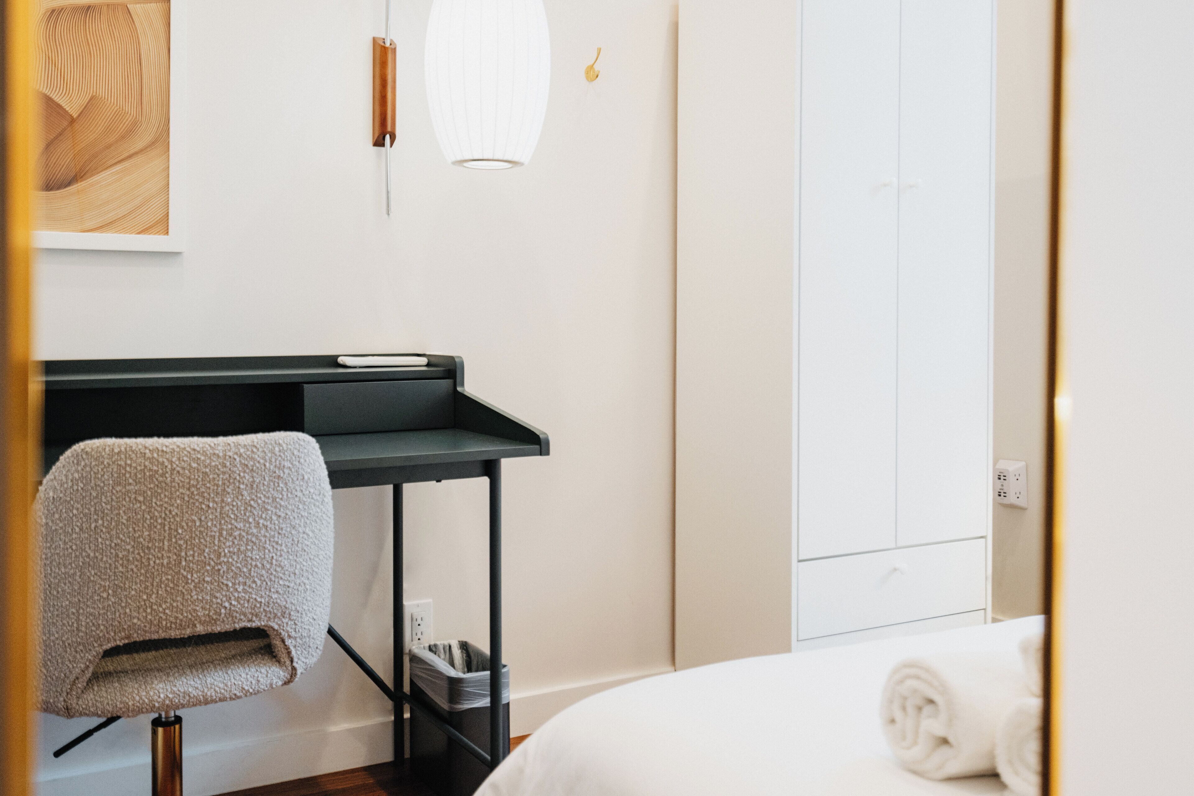 Una camera d'albergo con un letto, una scrivania con una sedia, un cestino e un punto luce a parete.