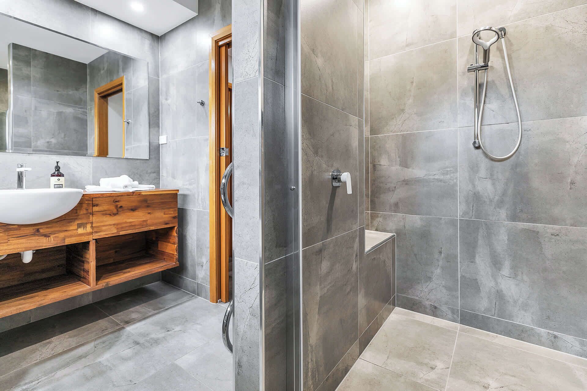 Een moderne badkamer met een houten wastafelmeubel, een grote spiegel en een inloopdouche met een handdouche.