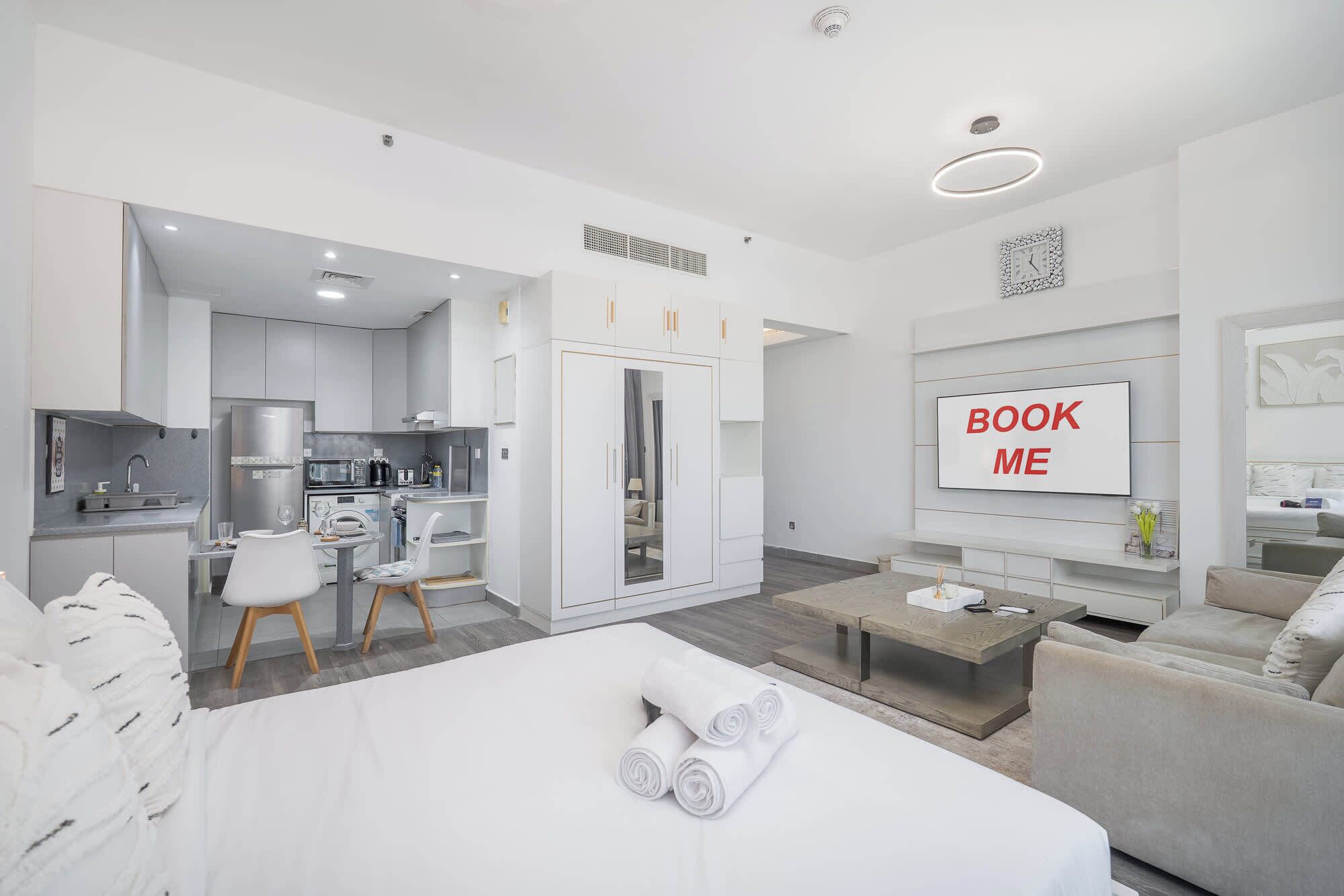 A modern hotel room with a kitchenette, a bed with rolled towels, a seating area with a sofa and armchair, and a TV screen with 'BOOK ME' text.