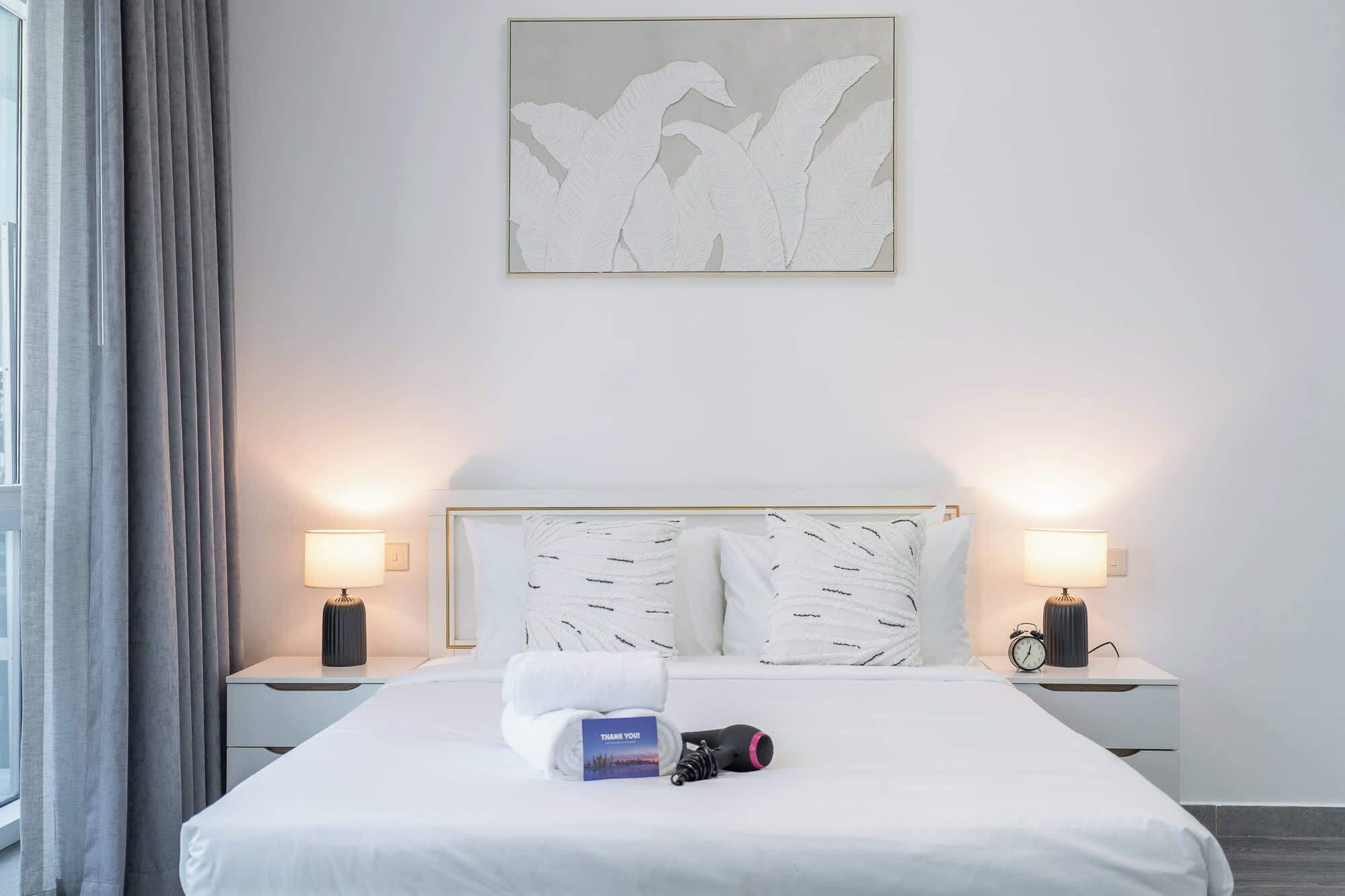 Ein Hotelzimmer mit einem großen Bett, zwei Nachttischlampen, einem Wandbild und einem Nachttisch mit einer Uhr und einer kleinen Lampe.