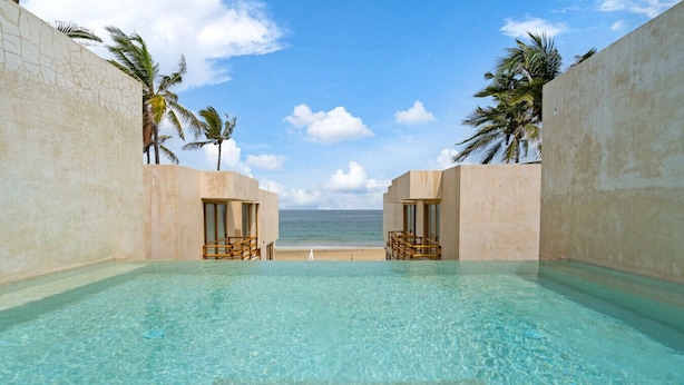Une piscine aux eaux cristallines, offrant une vue sur l’océan, entourée de palmiers et dotée d’une architecture simple.