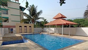 A swimming pool with tiled steps leading into it, surrounded by a low wall and a covered seating area. - Atithi Farm House (Kashid)