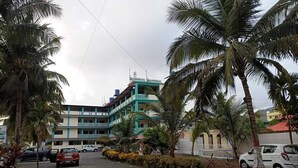 A building with a green and white facade, surrounded by palm trees and parked cars. - Atithi Farm House (Kashid)
