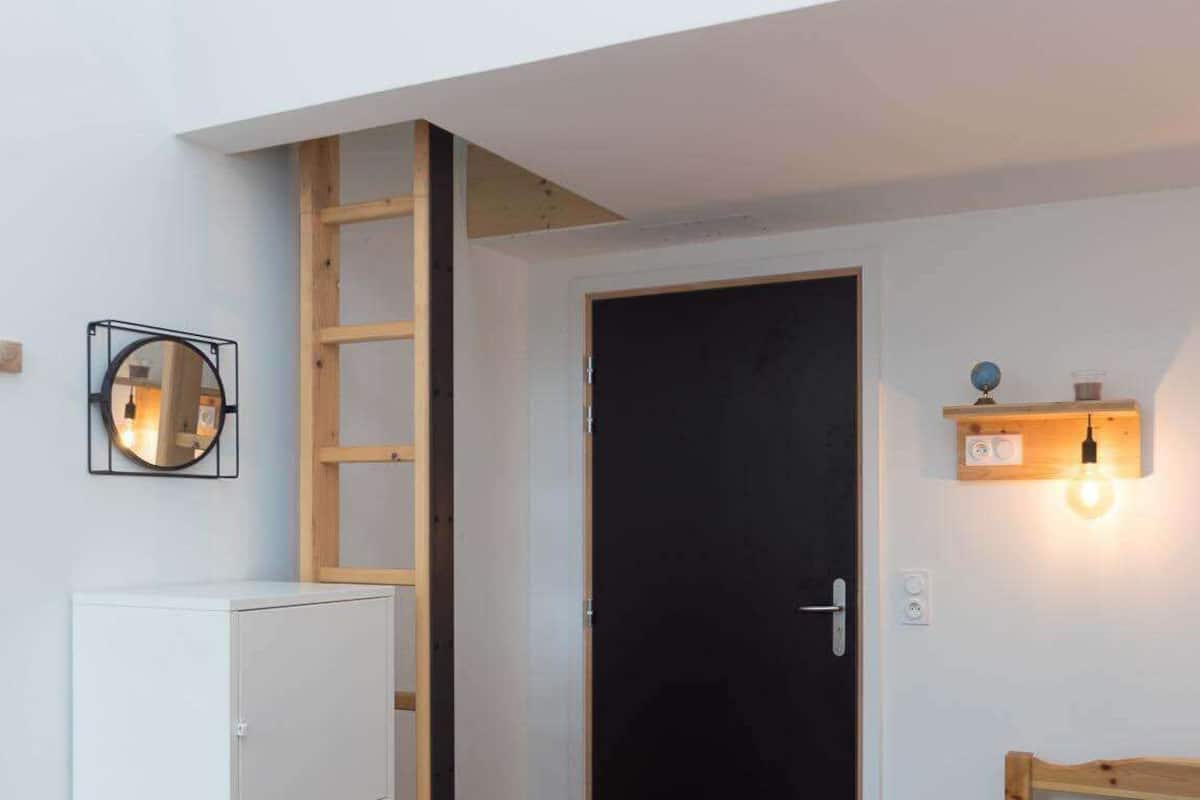 A room with a black door, a wooden ladder, a white cabinet, a round mirror, a wall-mounted light fixture, and a framed picture.