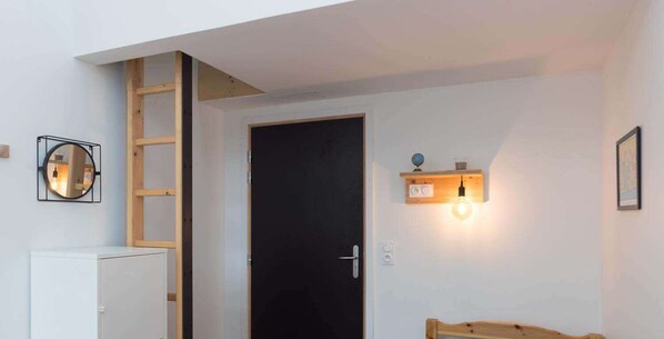 Ein Zimmer mit einer schwarzen Tür, einer Holzleiter, einem weißen Schrank, einem runden Spiegel, einer Wandleuchte und einem gerahmten Bild.