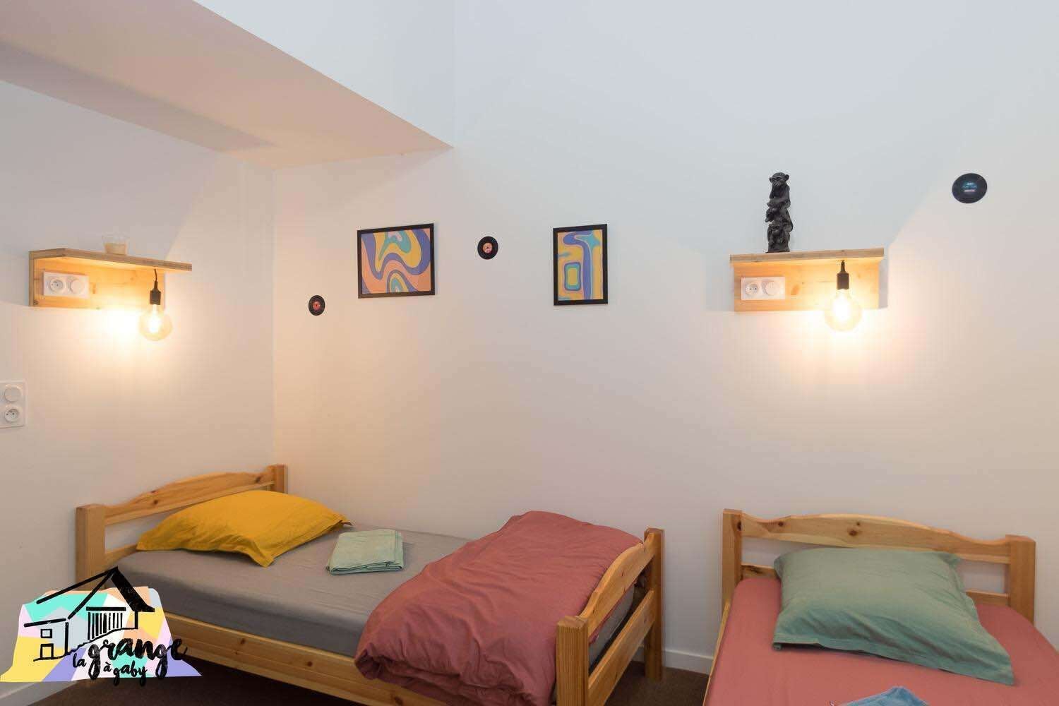 Une pièce avec deux lits en bois, chacun muni d’un oreiller et d’une couverture. On y trouve des appliques murales et deux œuvres d’art encadrées.
