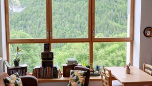 A room with a large window offering a forest view, a bookshelf, a small table, and a chair. - La Grange à Gaby (Engins)