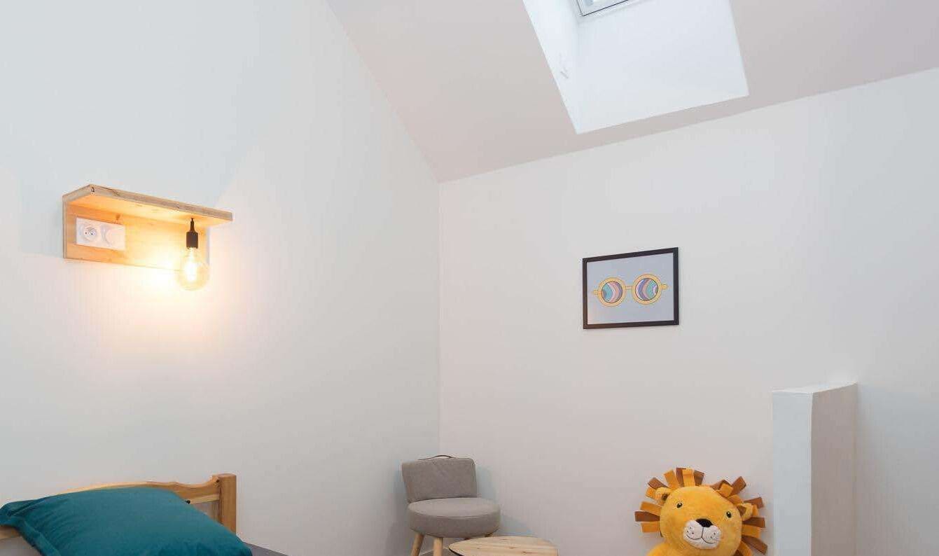 A bedroom with a skylight, a bedside lamp, a framed picture, a plush toy, and a small round table.