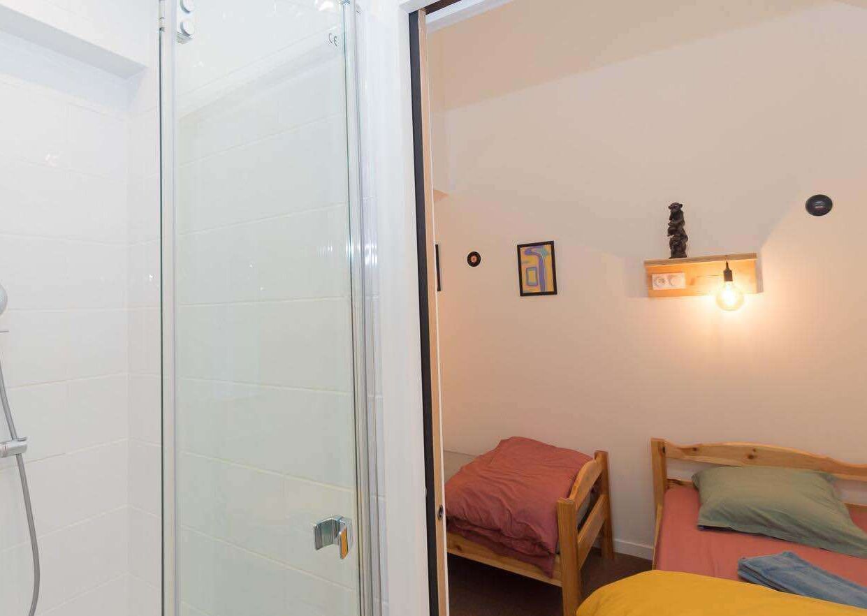 A small, compact room with a shower, a single bed with a wooden headboard, and a wall-mounted shelf with a light.