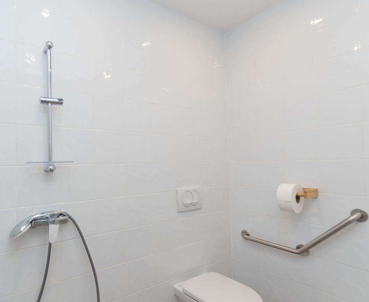 Een badkamer met witte tegels, een douche met handdouche, een toilet en een handgreep.