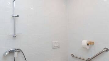 Een badkamer met witte tegels, een douche met handdouche, een toilet en een handgreep.