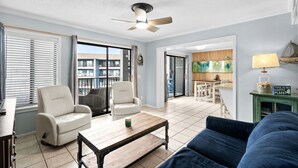 Een woonkamer met een plafondventilator, twee witte fauteuils, een blauwe bank, een houten salontafel en een keuken op de achtergrond.