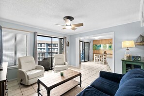 Ein Wohnzimmer mit Deckenventilator, zwei weiße Sessel, ein blaues Sofa, einem Holztisch und einer Küche im Hintergrund.