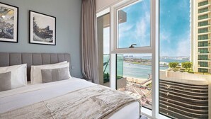 Una moderna camera d'albergo con un letto grande, due opere d'arte incorniciate, vista sulla città attraverso ampie finestre e un balcone con vista su un fiume e degli edifici.