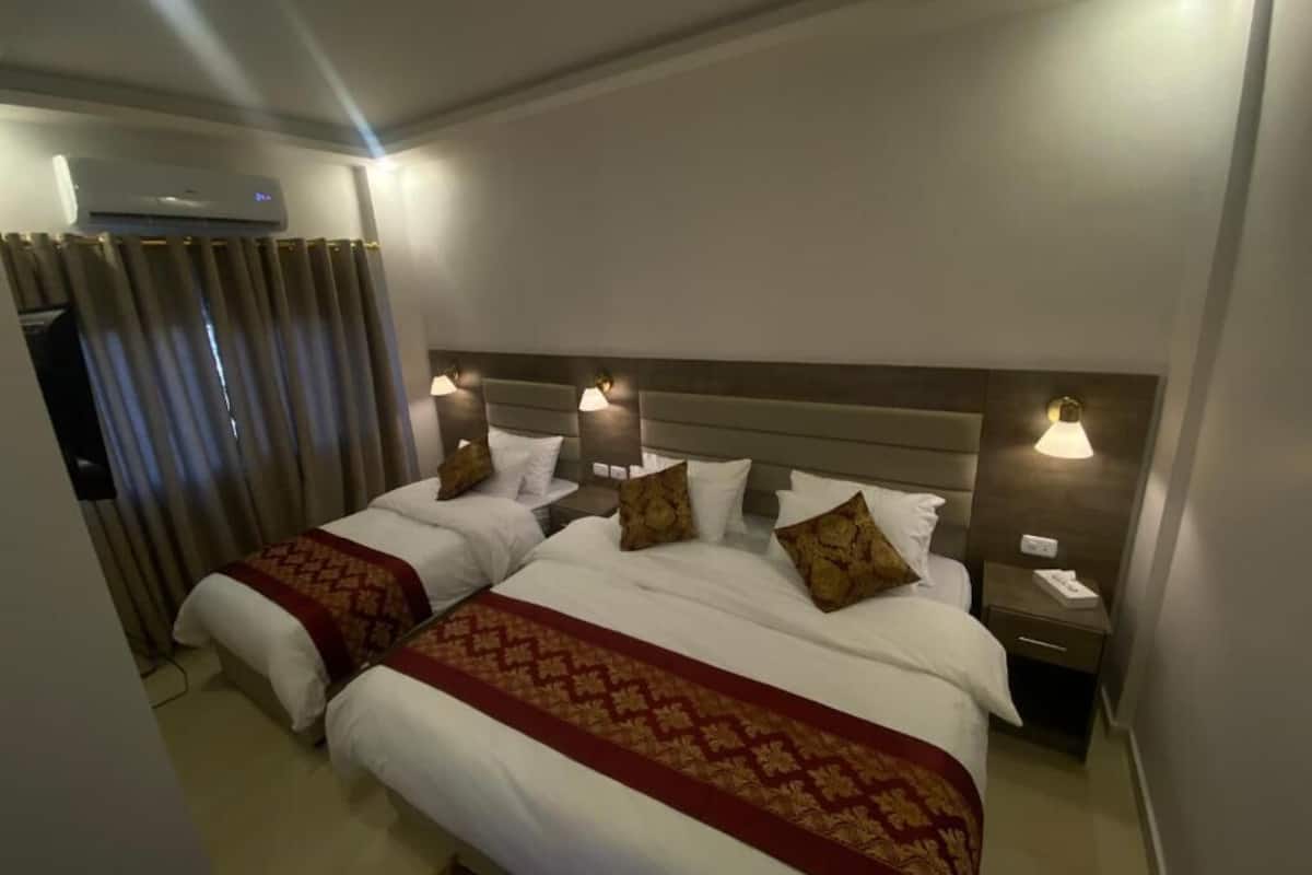 A hotel room with two beds, each with a headboard, bedside lamps, and a red patterned bedspread.