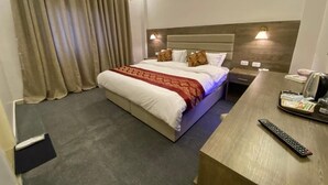 A hotel room with a bed, curtains, a bedside table with a remote control, and an air conditioning unit. - Petra Oleander Hotel (Wadi Musa)