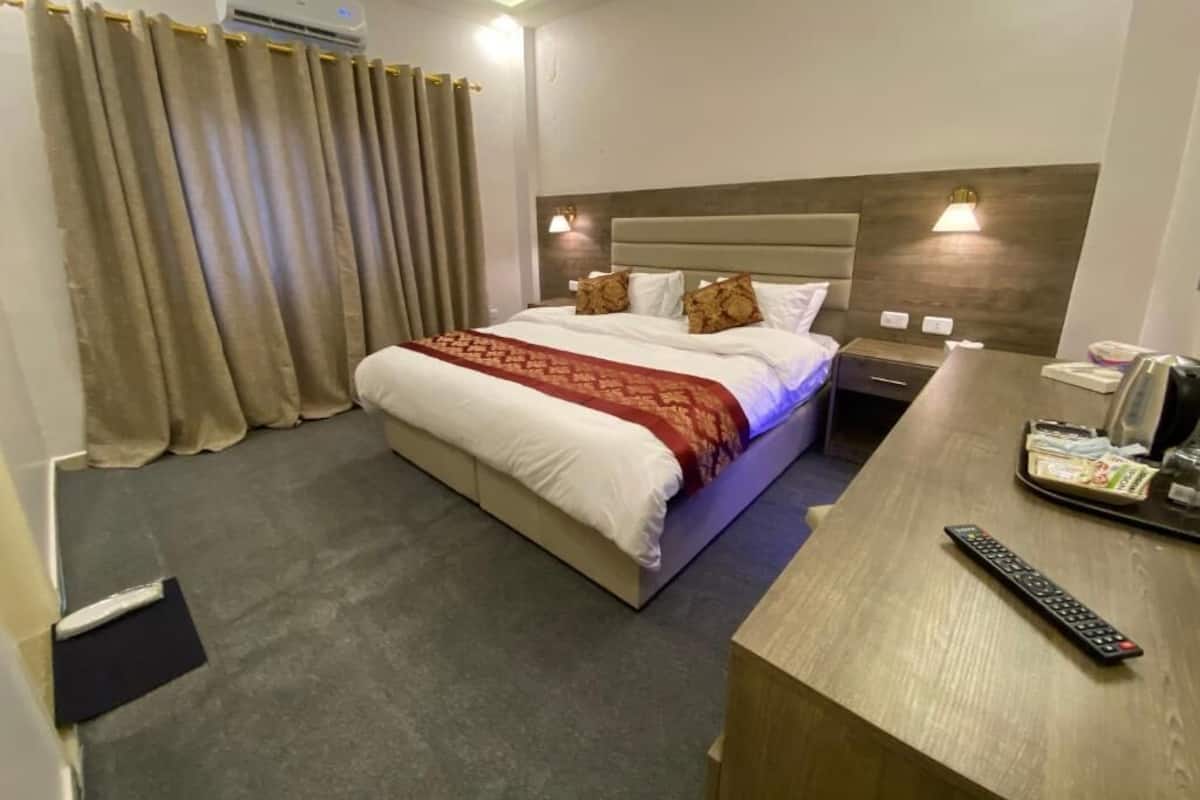 A hotel room with a bed, curtains, a bedside table with a remote control, and an air conditioning unit.