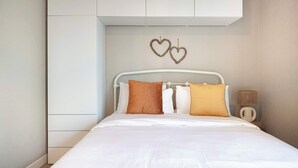 A neatly made bed with white and brown bedding, a white metal headboard, and two heart-shaped decorations on the wall.