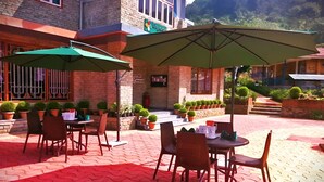 An outdoor patio with tables and chairs, green umbrellas, potted plants, and a brick building in the background. - Khachoe Residency, Ravangla (Namchi)