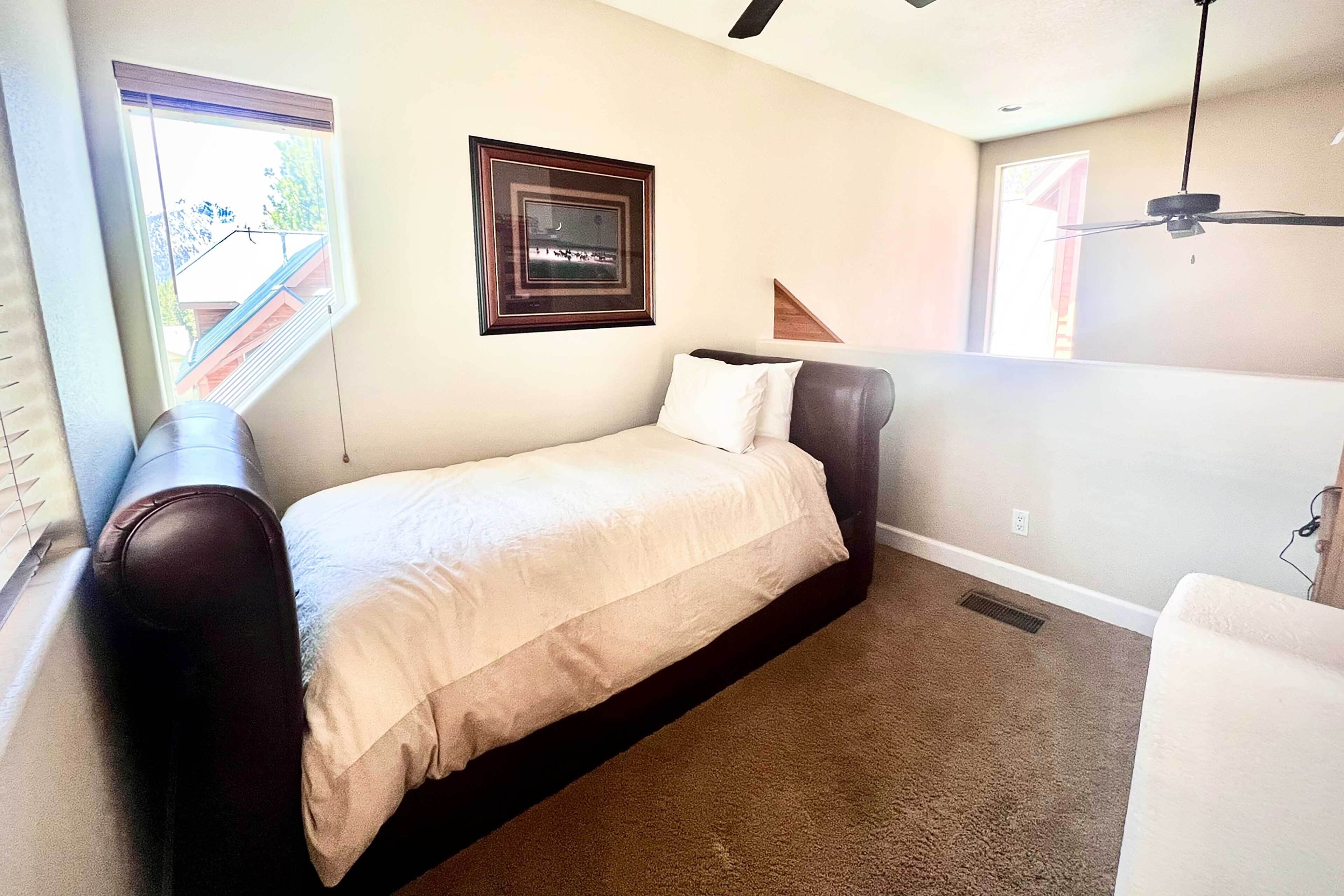 A small, single-bed room with a window, a framed picture on the wall, a ceiling fan, and a carpeted floor.