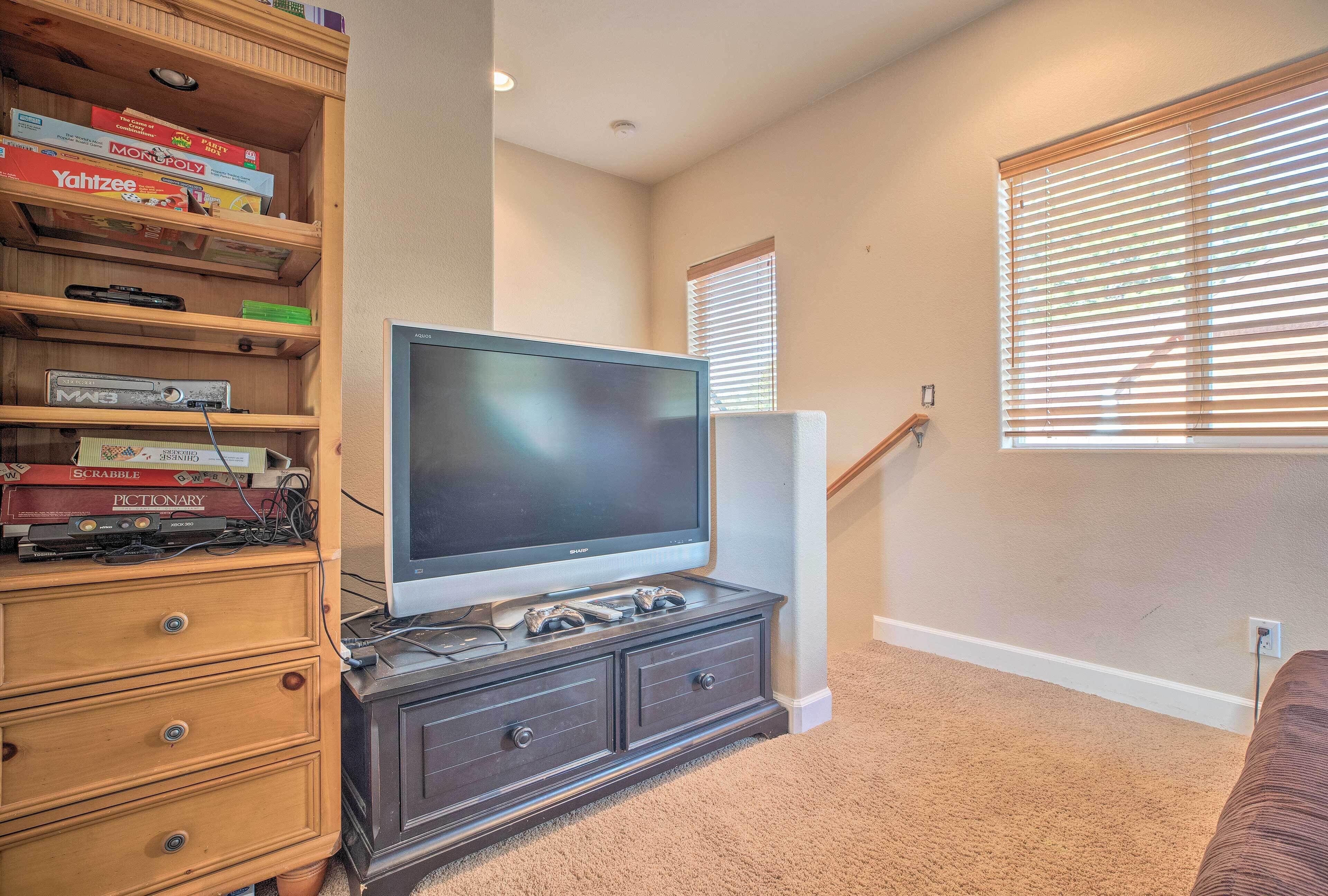 A living room with a flat-screen TV on a wooden cabinet, a bookshelf with board games, and a staircase with a window with blinds.