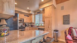 A kitchen with wooden cabinets, a granite countertop, a stainless steel refrigerator, a microwave, a stove, and a sink. There is a trash can and a chair in the corner. - Mammoth Lakes Townhome - 10-min Walk to Village! (Mammoth Lakes)