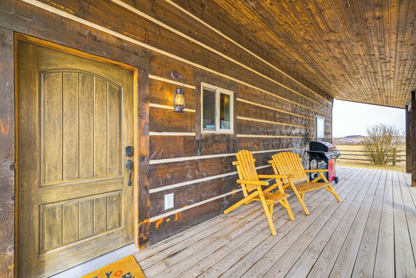 Egy fából készült, verandás ház, két Adirondack-stílusú székkel, egy grillsütővel és egy bejárati szőnyeggel ellátott ajtóval.