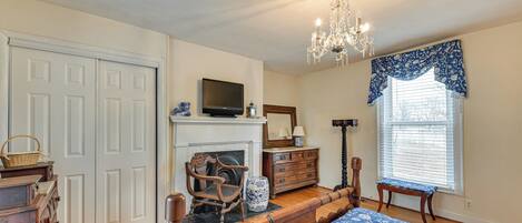A bedroom with a wooden bed, a television on a mantel, a fireplace, a chandelier, and a window with blinds.
