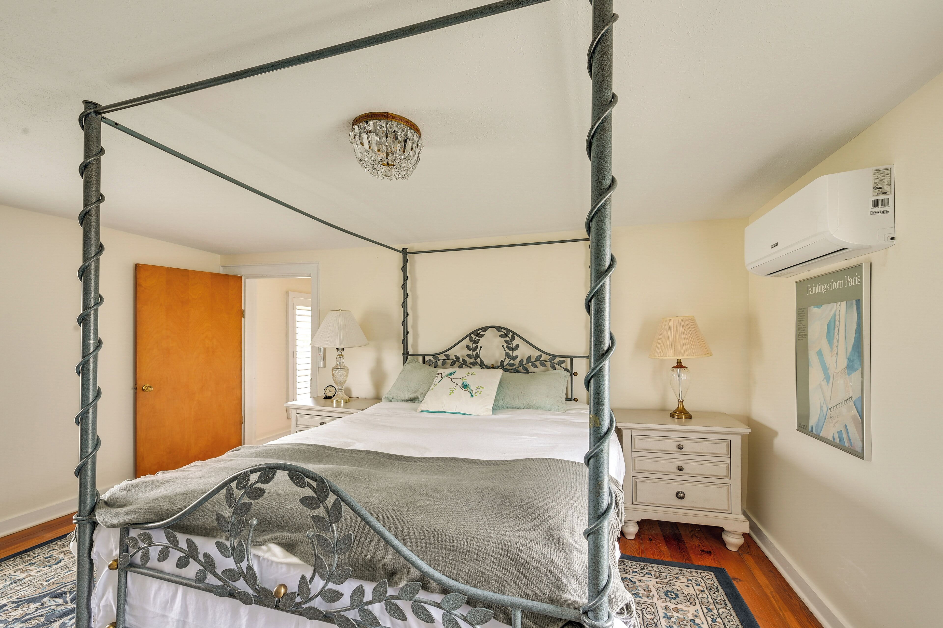 A four-poster bed with a leaf-patterned metal frame, a chandelier, a bedside table with a lamp, and a framed picture on the wall.