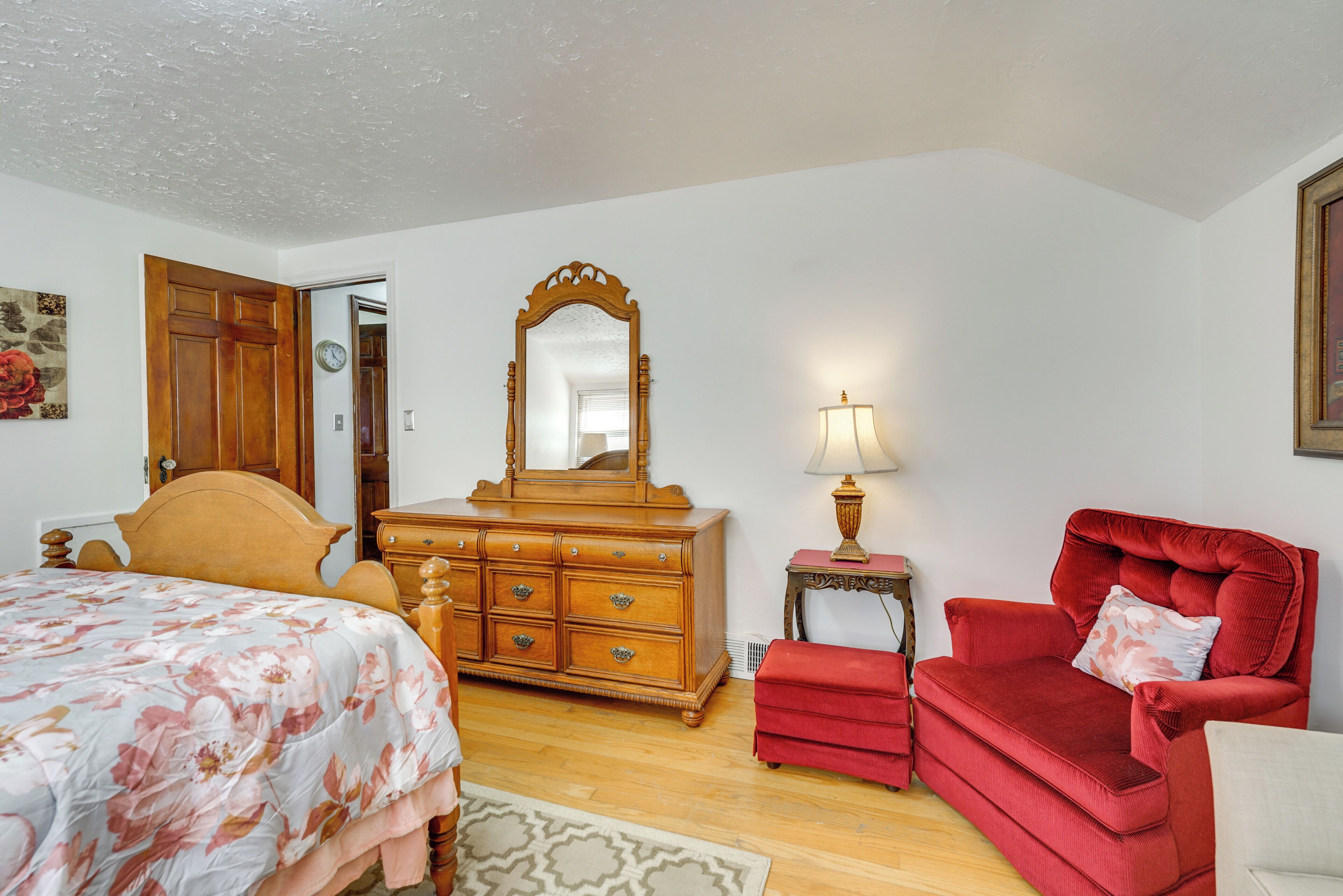 A bedroom with a bed, dresser, mirror, red armchair, and a small table with a lamp.