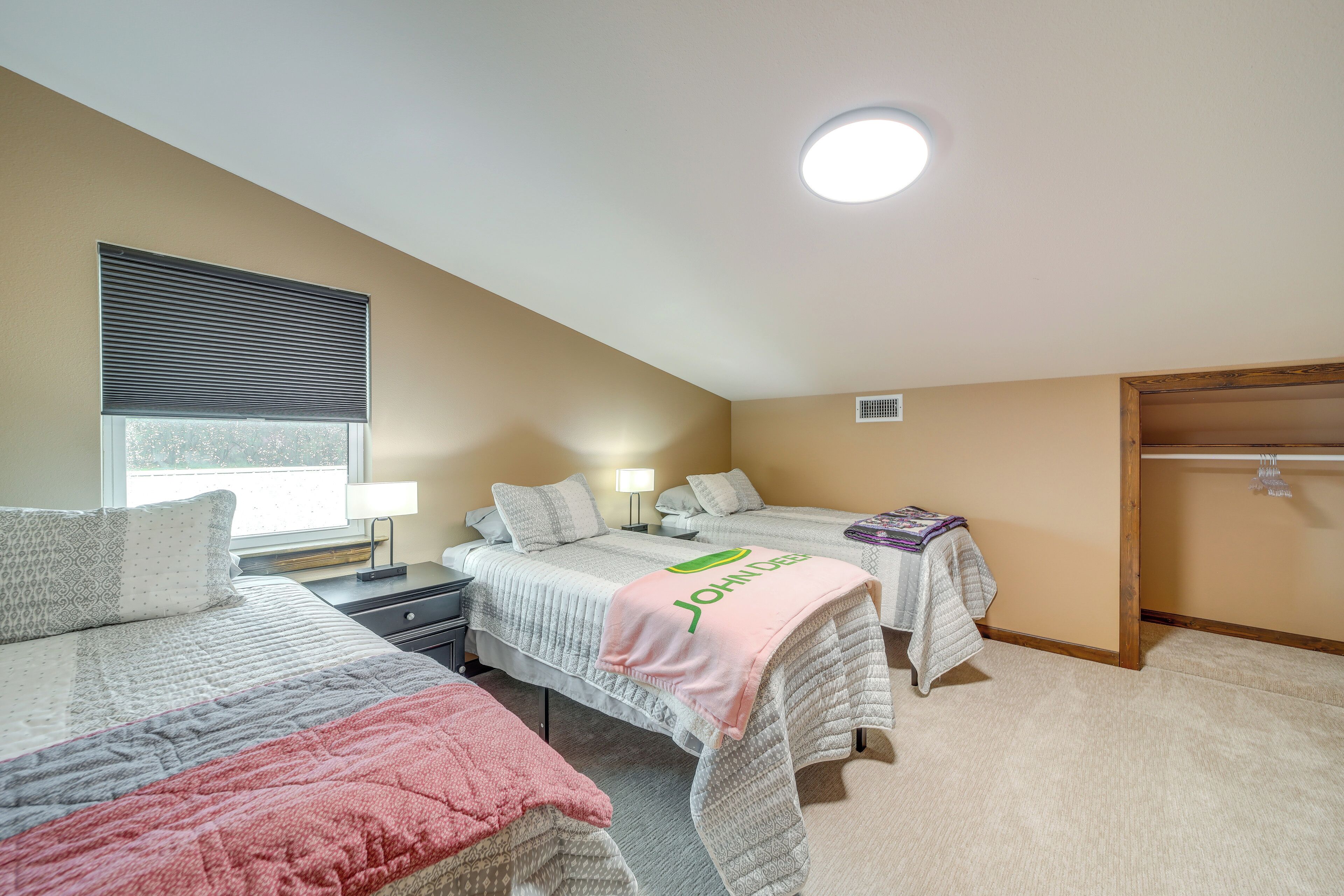 A bedroom with two beds, a window with blinds, a bedside table with a lamp, and a closet with a hanging rod.