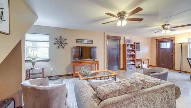 A spacious living room with a sofa, armchairs, a coffee table, and a television. There are ceiling fans, a bookshelf, and a window with blinds.