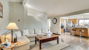 Ein modernes Wohnzimmer mit Sofa, Couchtisch und Lampe. Im Hintergrund ist eine Küche mit Kühlschrank und Barhocker zu sehen.