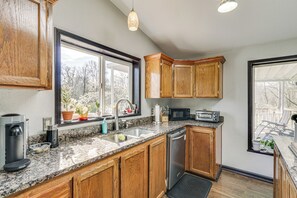 A kitchen with wooden cabinets, granite countertops, a built-in dishwasher, a microwave, and a large window with a view of trees.