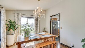 Ein Essbereich mit Holztisch, Bänken, einer Chandelüster, Topfpflanzen und Blick auf eine Küche und einen Kühlschrank.