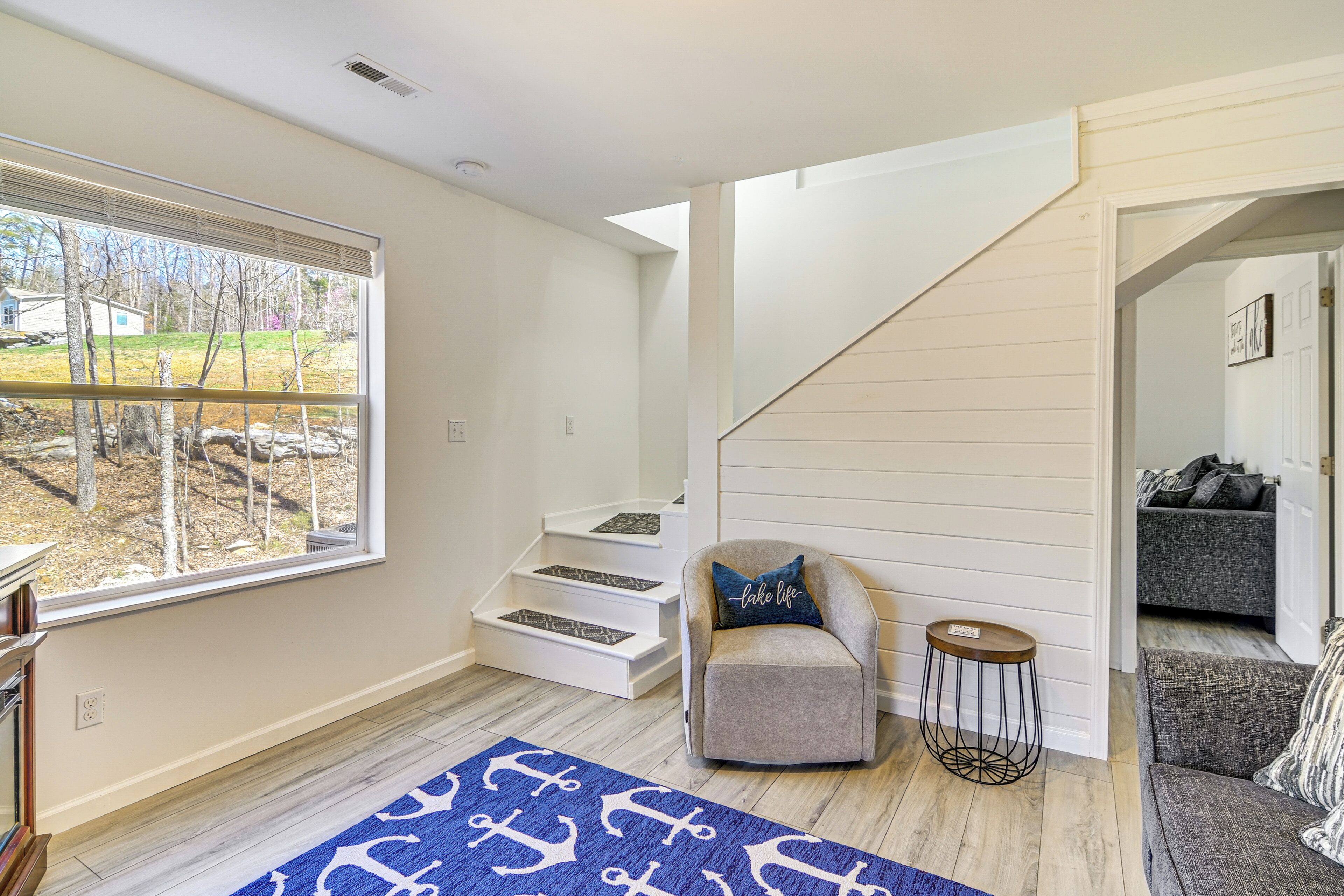 A modern living room with a staircase, a grey armchair, a round side table, and a blue rug with anchor designs.