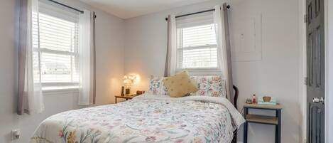 Une chambre à coucher avec une couvre-lit à motifs floraux, deux fenêtres avec des rideaux, une lampe de chevet et une petite table de nuit en bois.