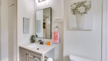 Un baño con un lavabo blanco, un espejo, un perchero para toallas, un inodoro y una obra de arte enmarcada en la pared.