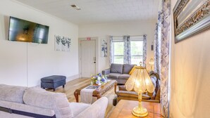 A living room with a grey sofa, a small table with a lamp, and a TV mounted on the wall. - Half-mi to Dtwn Lufkin: Charming & Spacious Home! (Lufkin)