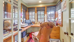 Ein gut beleuchtetes Büro mit Schreibtisch, Computer und verschiedenen Büromaterialien. Ein Fenster mit Jalousien, ein Stuhl und ein Bücherregal sind ebenfalls vorhanden.