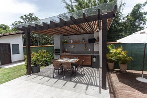 An outdoor kitchen area with a pergola, dining table, chairs, and potted plants. - Bz10 Casa 4 suítes em condomínio clube (Buzios)