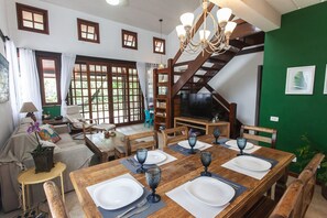 A dining area with a wooden table set for four, a staircase, and a television in the background. - Bz10 Casa 4 suítes em condomínio clube (Buzios)