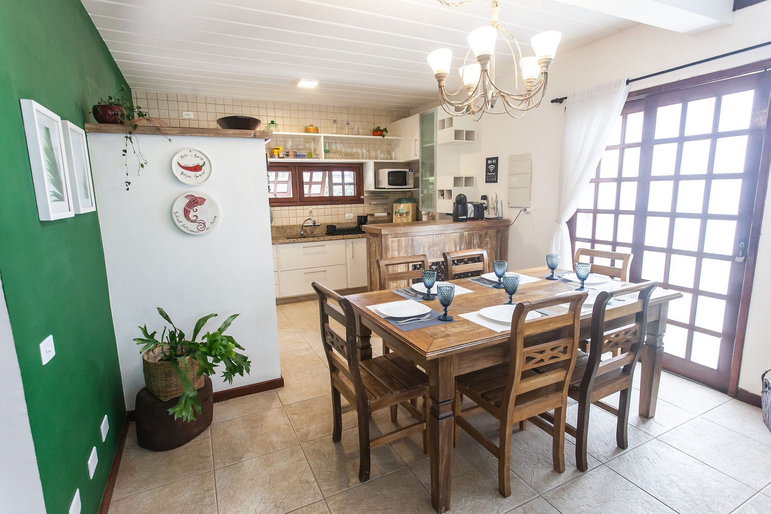 A dining area with a wooden table and chairs, a chandelier, and a view of the kitchen in the background.