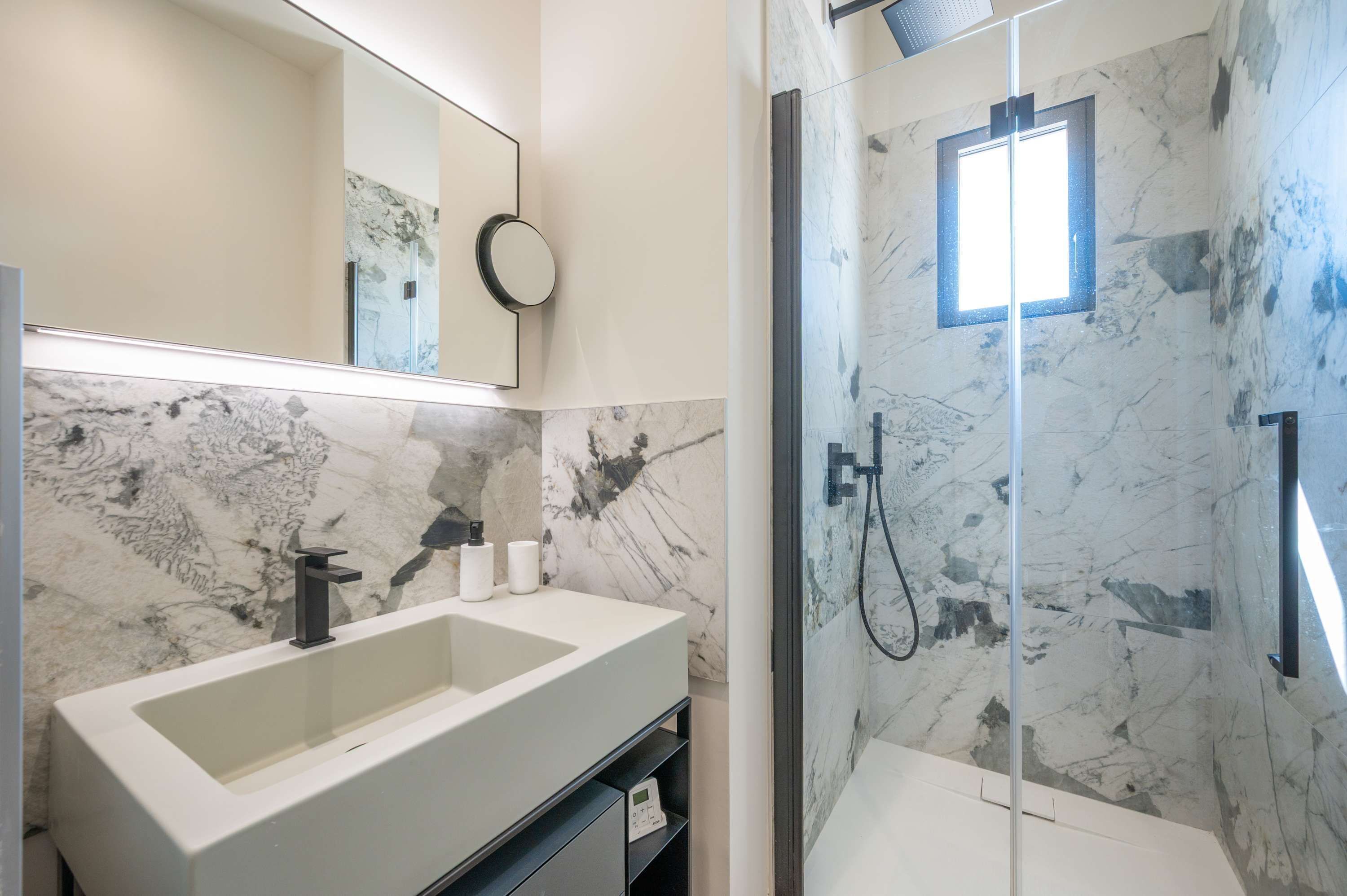A bathroom with a double sink, a mirror, a handheld showerhead, and a glass-enclosed shower area with a marble wall.