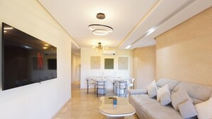 Un salon moderne avec un canapé, une table basse et un téléviseur fixé au mur. On aperçoit à l’arrière-plan un coin repas avec une table et des chaises.